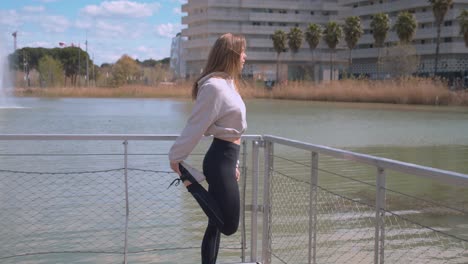 a girl in a tracksuit, standing on a bridge, pulling her legs back with her hands for doing exercises