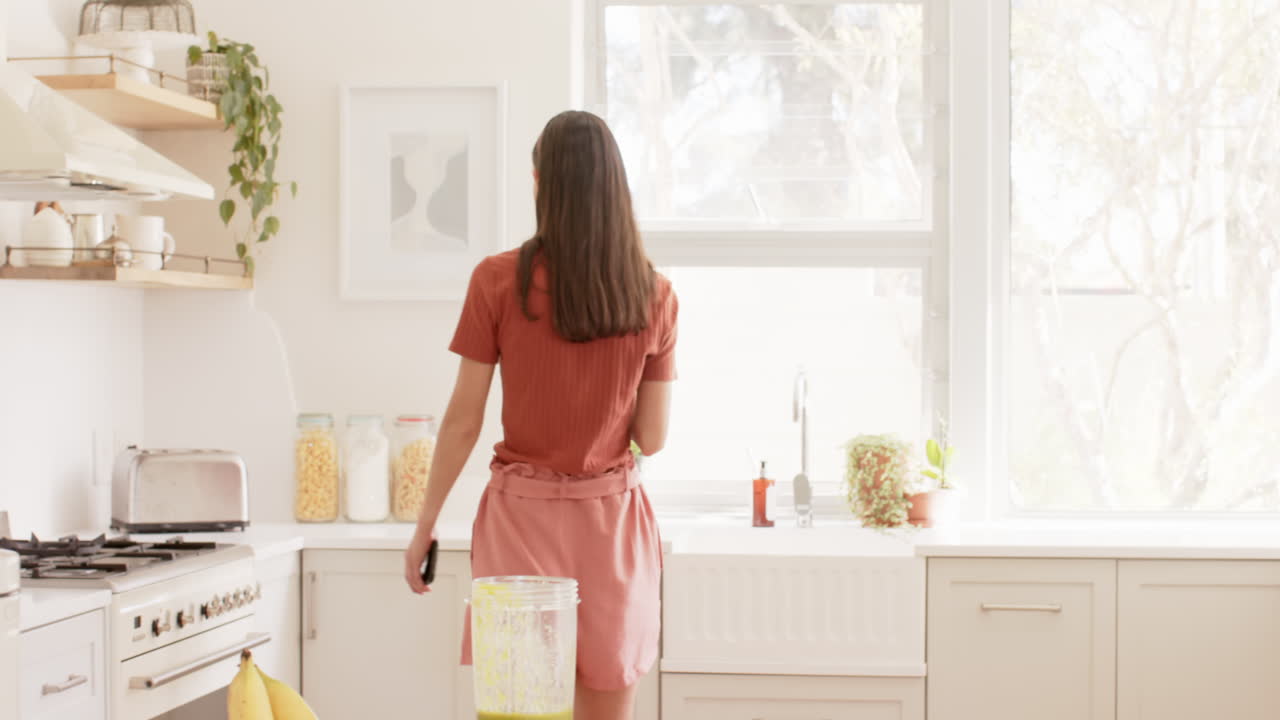 Smiling woman in kitchen using smartphone while preparing healthy smoothie