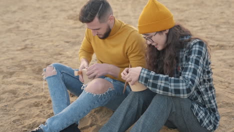 jóvenes amigos hombres y mujeres comiendo comida para llevar sentados en la playa.