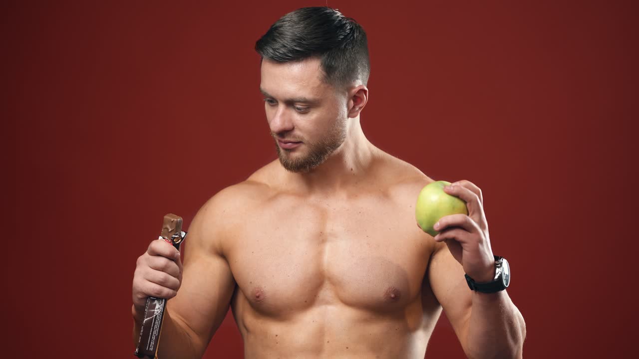 Portrait of a athletic man choosing between a fresh apple and a chocolate. Healthy food. Dieting concept. Half lenght video.