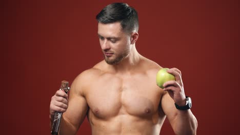portrait of a athletic man choosing between a fresh apple and a chocolate. healthy food. dieting concept. half lenght video.