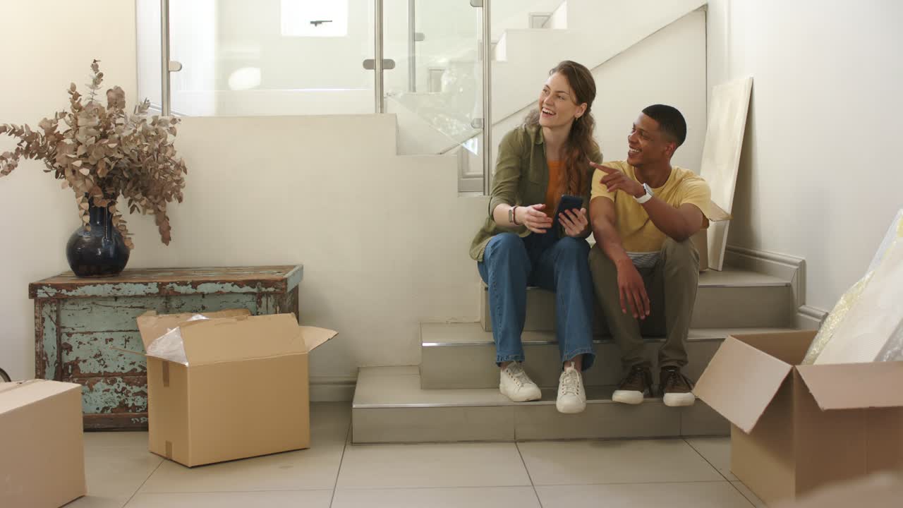 Un couple est assis sur un palier d'escalier au milieu de cartons de déménagement, l'homme pointant du doigt tandis que la femme vérifie un appareil lors du déménagement.