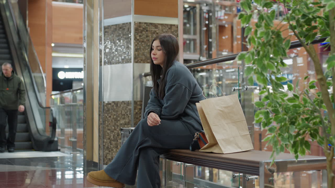 Young female shopper stands calmly in modern shopping mall after shopping, looking thoughtful while people descend escalator behind her, surrounded by shops, plants, and bright overhead lights