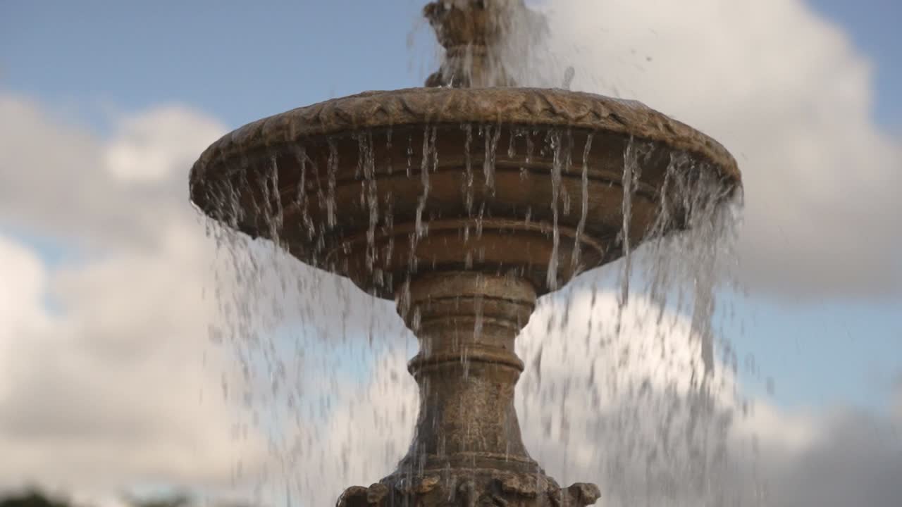 L'acqua sgorga da una fontana in un parco. Le persone passeggiano e si godono la giornata di sole. Il cielo è parzialmente nuvoloso. È un momento vivace in uno spazio pubblico circondato dal verde.