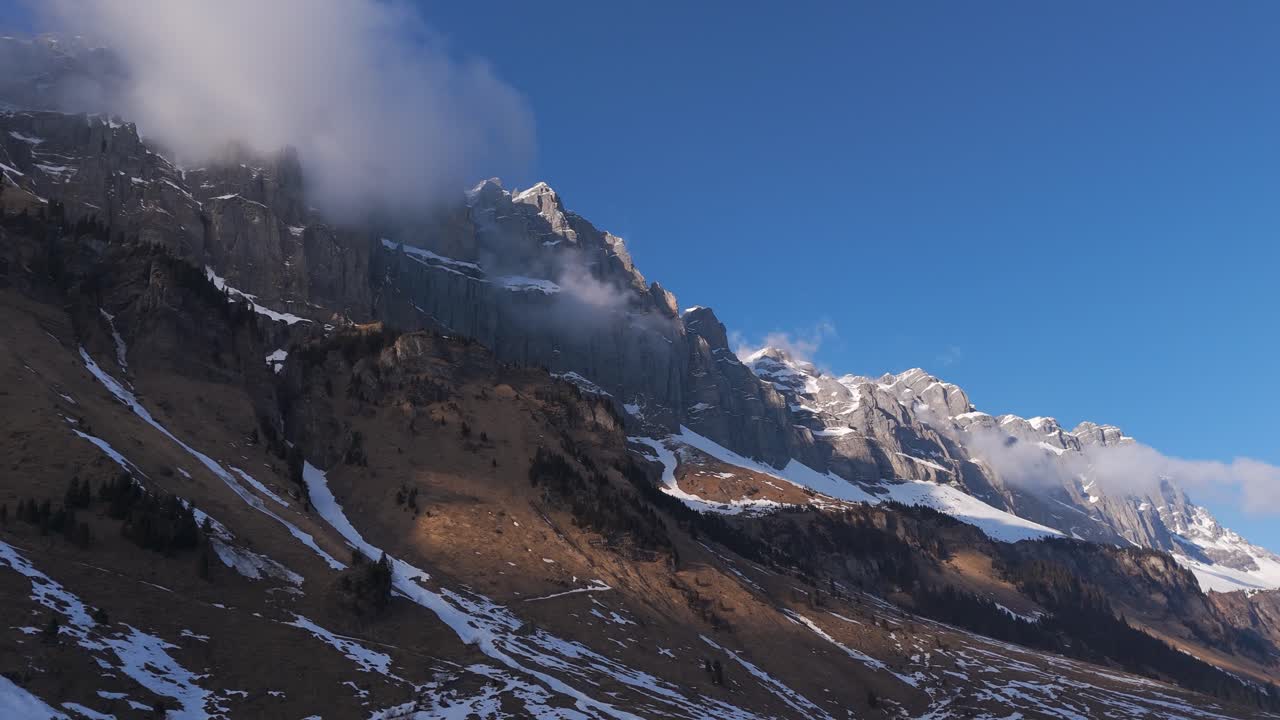 Drone flight high in the mountains. The mountain ranges and peaks are covered in snow. Clouds hang over the mountaintops. The sun shines on the massive mountains, and the sky is blue.