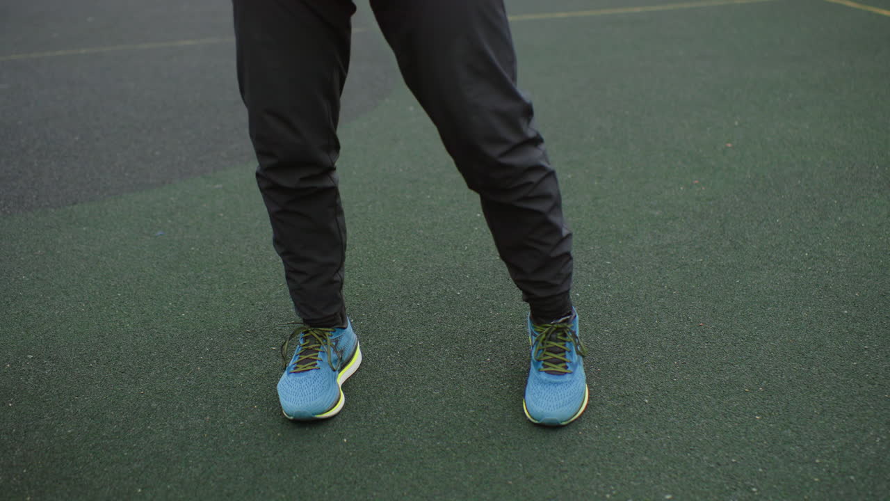 Piedi concentrati che si esercitano in esercizi di basket all'aperto, scarpe da ginnastica blu e pantaloni da jogging neri, gioco di gambe lento e preciso con palleggio controllato, texture verde del campo in gomma visibile, ripetizione disciplinata per l'abilità