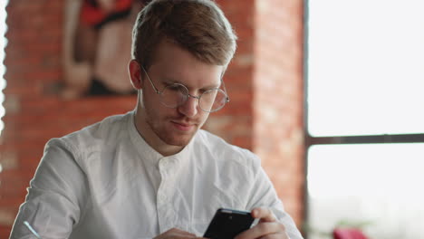 Un-Hombre-Guapo-Con-Gafas-Está-Charlando-En-Una-Aplicación-En-Un-Teléfono-Inteligente-Sentado-Solo-En-Un-Café-Viendo-Noticias-En-Las-Redes-Sociales