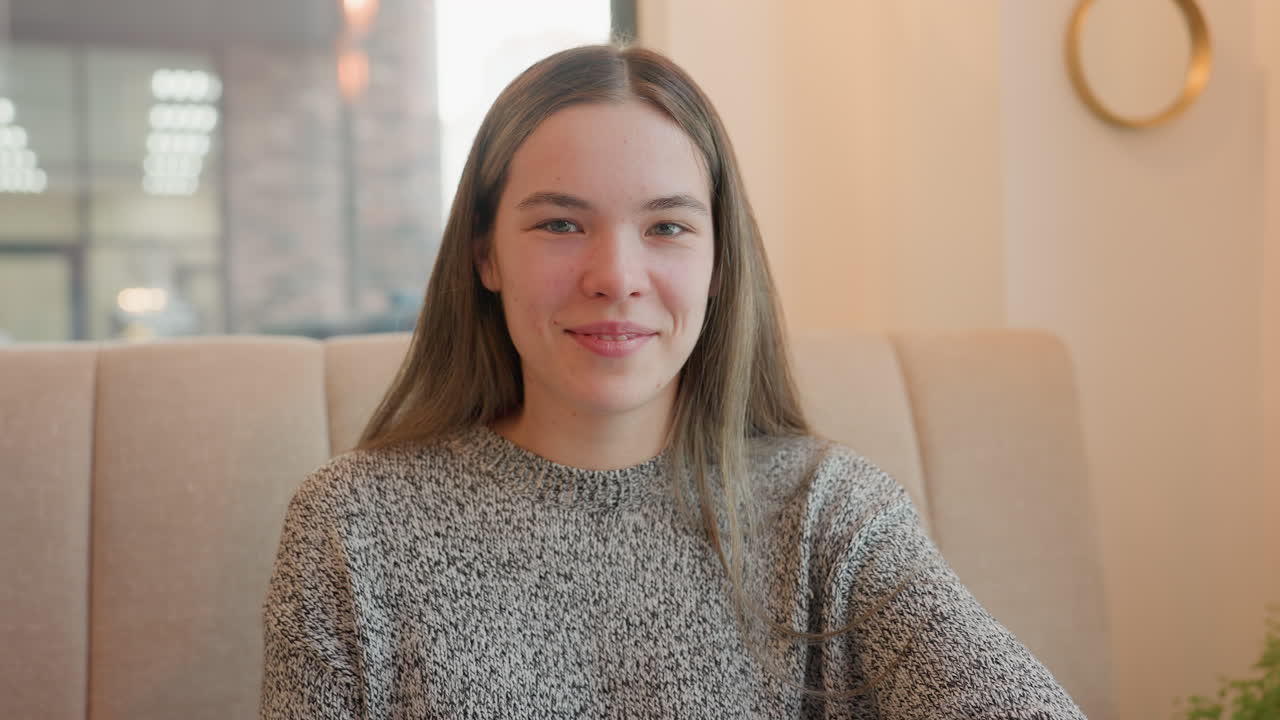 Cheerful young woman smiling and sitting in coffee bar enjoying her time while watching cars pass through street visible through transparent window, cozy atmosphere, and daylight outside