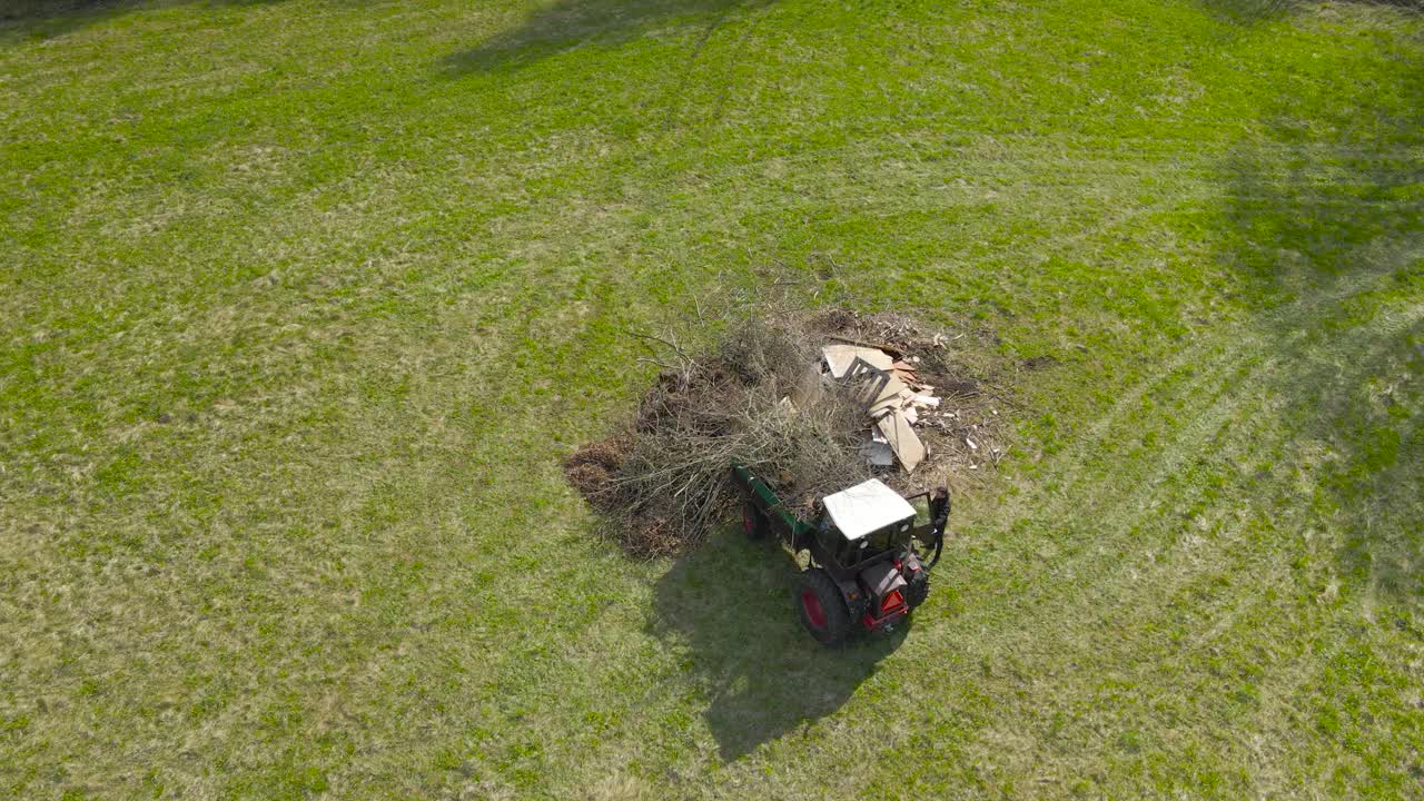 Aerial drone footage flying over an old vintage retro tractor that has its loader full of cut tree branches and garden waste in a green grassy field during a summer sunny day while a man is standing.