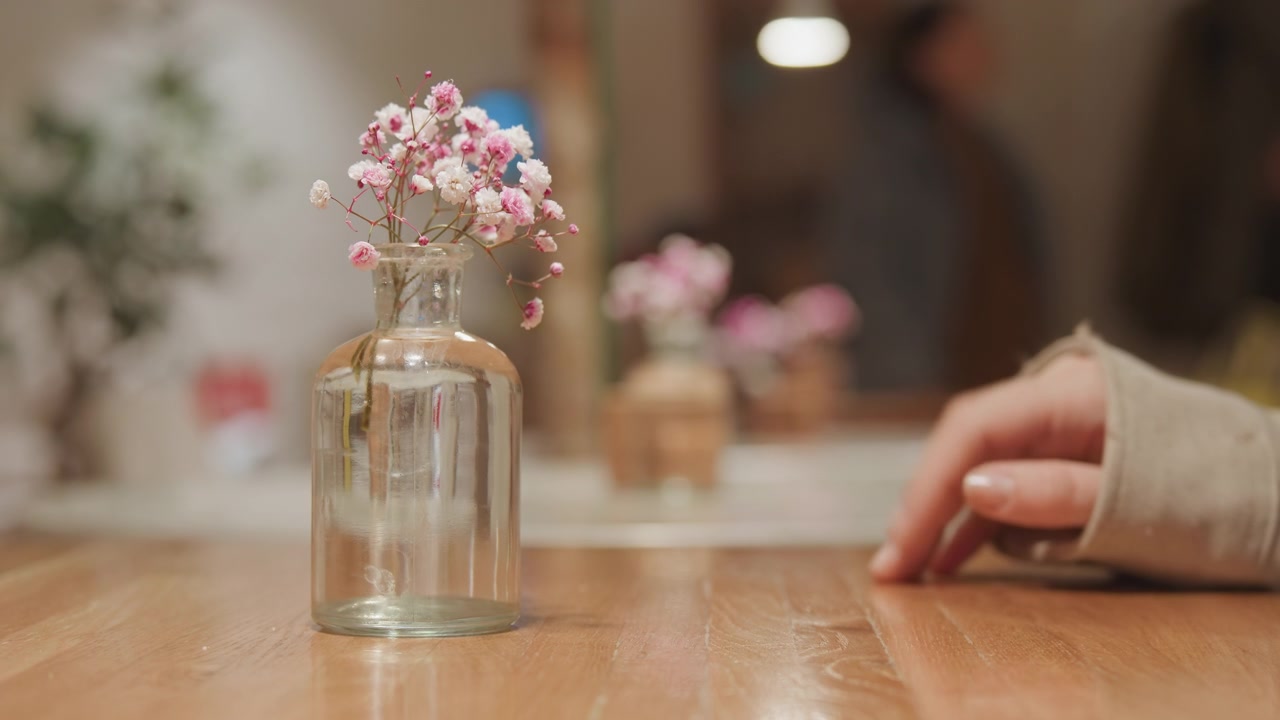 Gros plan d'un vase en verre transparent avec des fleurs roses sur une table en bois. Une main en manche longue s'approche d'une bouteille vide puis se rétracte, avec un éclairage doux et un arrière-plan flou présentant un élément similaire