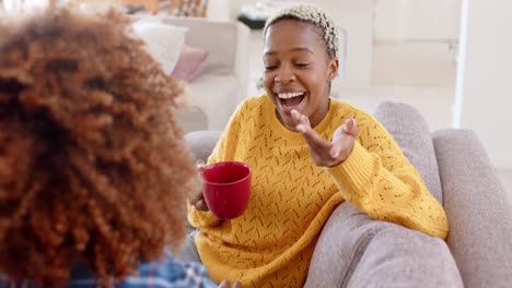 Happy-diverse-female-lesbian-couple-talking-and-drinking-coffee-in-living-room-in-slow-motion