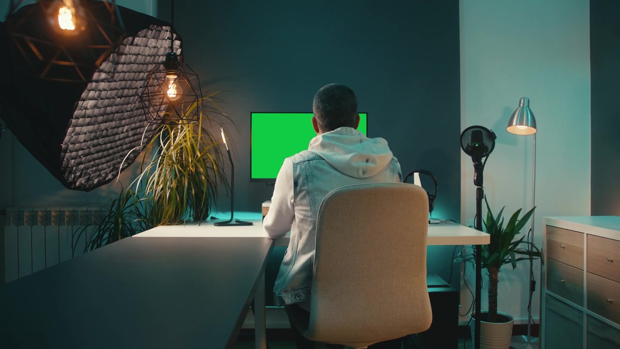 Teal and orange home office studio with modern tech. Young man in denim vest works at stylish desk with desktop computer and microphone. Content creation or social media concepts. Chroma key monitor.