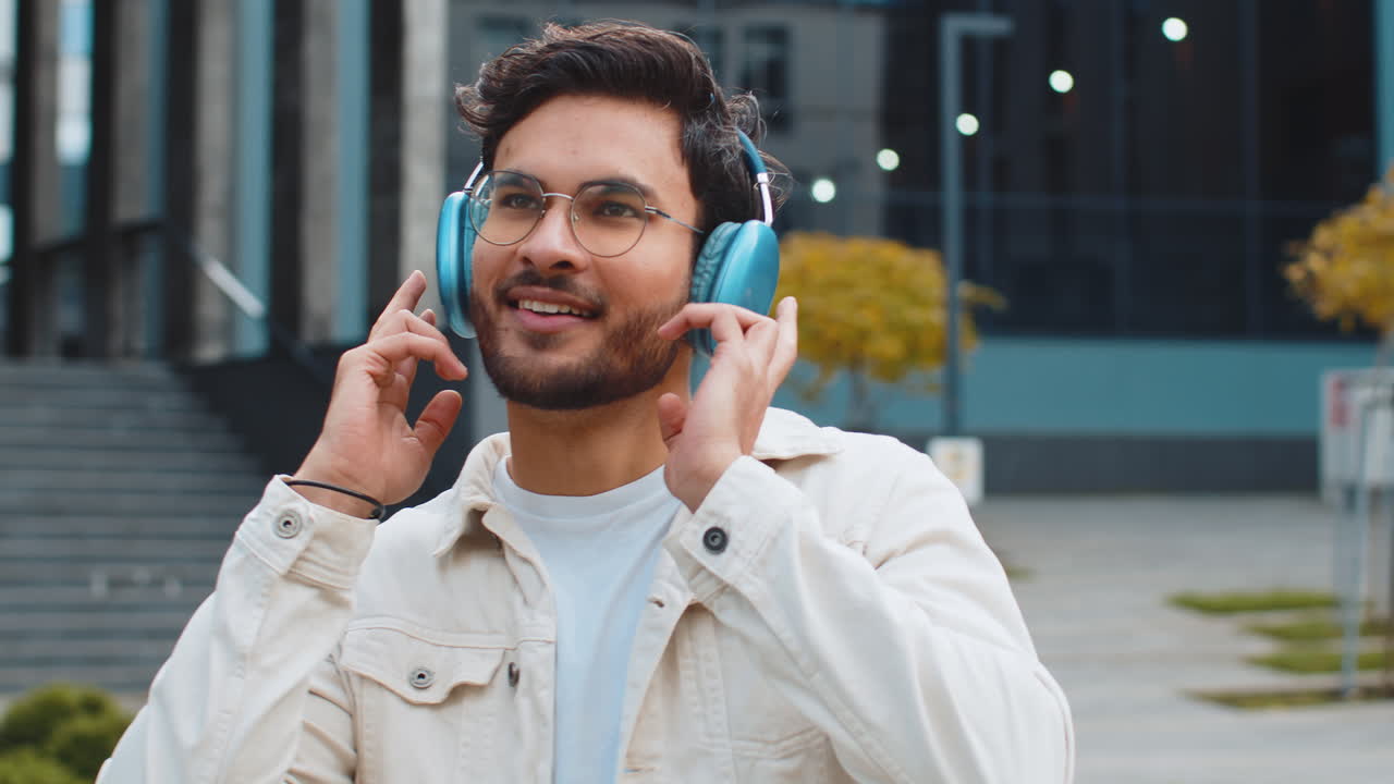 Happy indian man in headphones choosing listening music dancing standing outdoors city street