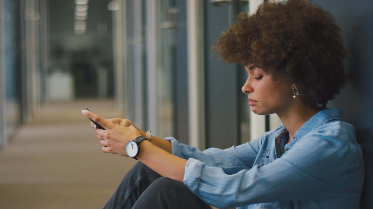 Stressed Young Businesswoman Sitting On Floor In Corridor Of Modern Office With Phone