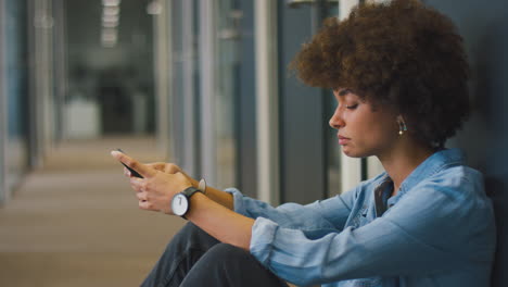 Gestresste-Junge-Geschäftsfrau-Sitzt-Mit-Telefon-Auf-Dem-Boden-Im-Flur-Eines-Modernen-Büros