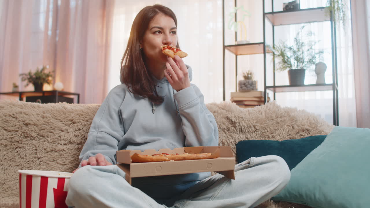 Giovane donna che mangia una deliziosa pizza e popcorn (cibo spazzatura) seduta sul divano a guardare la TV a casa