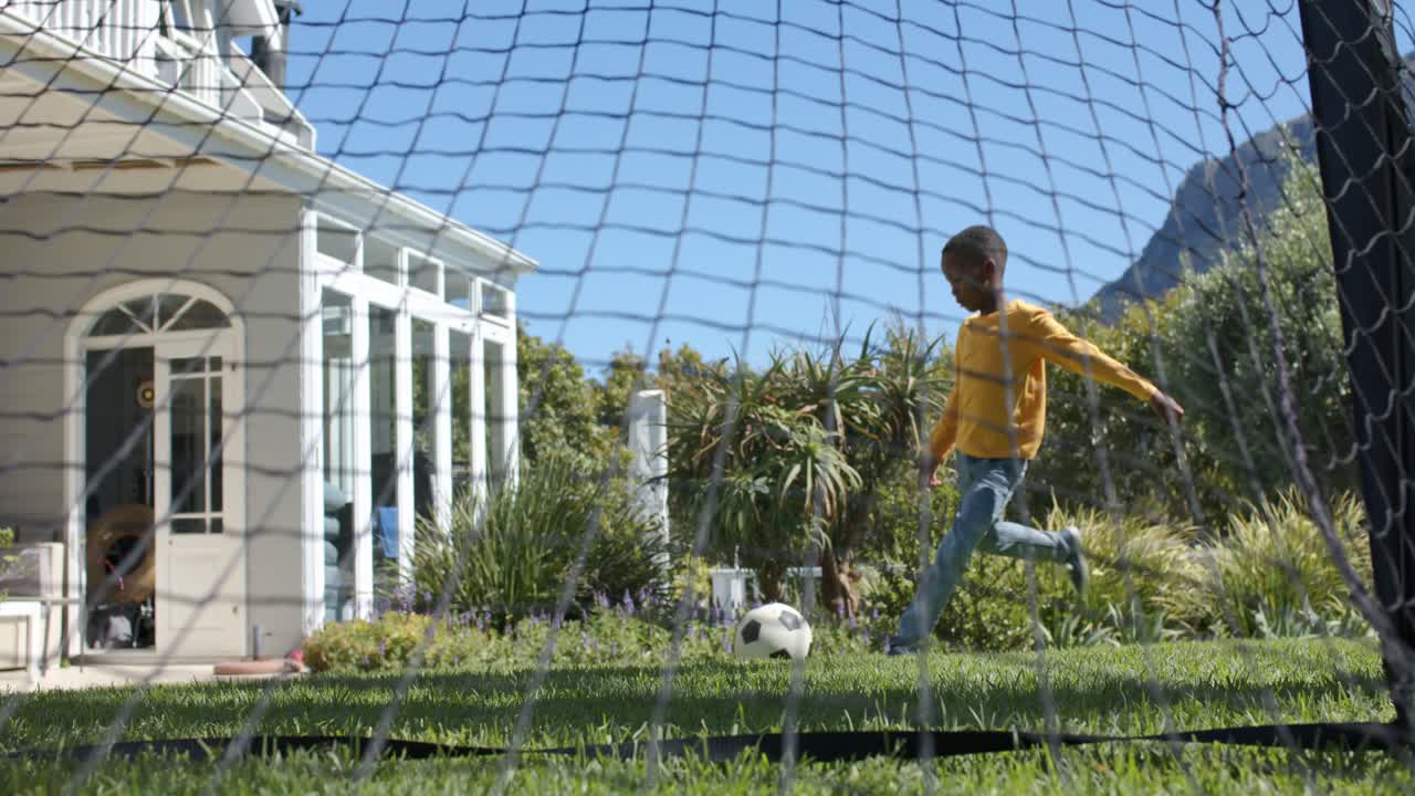 African American dad watching son nudging, kicking soccer ball via goal net in backyard practicing