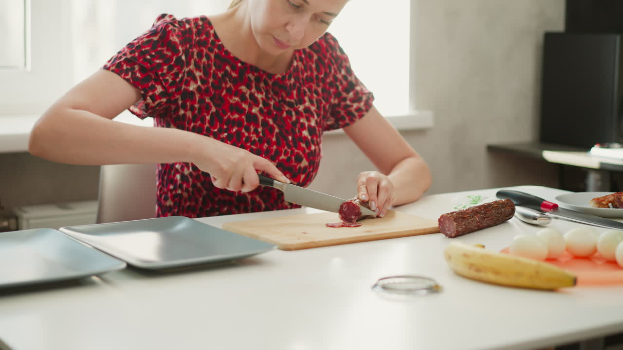 Chef slicing small pieces of cured sausage on wooden chopping board over white table by bright window light showcasing careful culinary technique and textured meat closeup