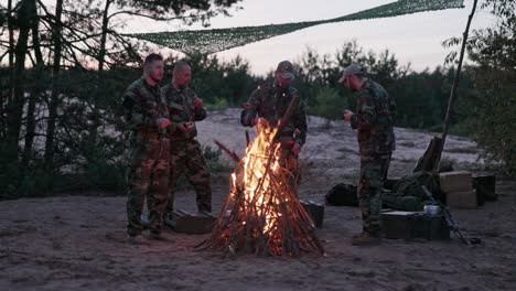 four men in camouflage uniforms stand around a hearth with a bonfire after sunset, frying sausages on sticks, uniformed men resting after duty, field exercises