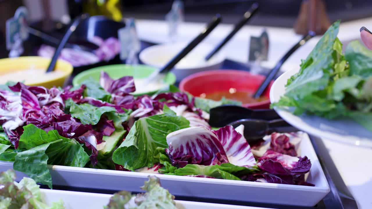 Primo piano di una persona in un ristorante che usa le pinze e il mestolo a un colorato buffet di insalate, ripresa a mano