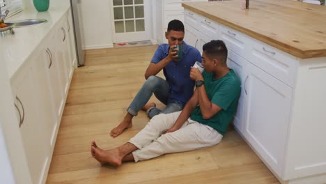happy mixed race gay male couple sitting on kitchen floor talking and drinking coffee