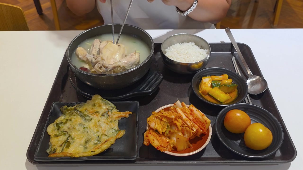Nampan makanan Korea lengkap menampilkan sup ayam samgyetang dengan nasi, kimchi, acar timun, panekuk makanan laut, dan hidangan sampingan banchan plum acar