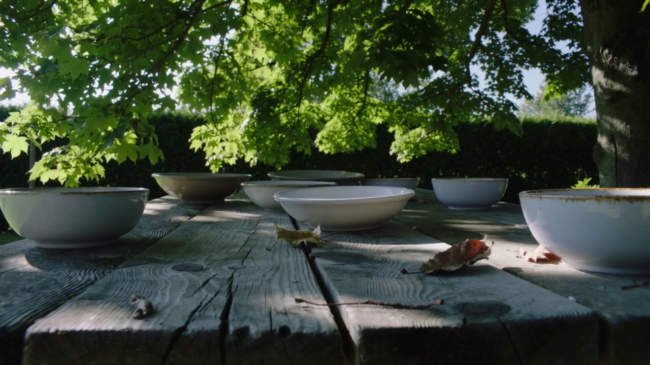 Une légère brise déplace des feuilles séchées, faisant danser le soleil tacheté sur une table en bois sous un arbre, avec des bols en céramique.