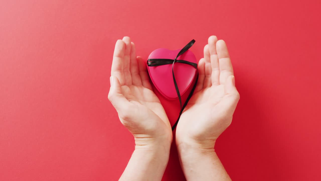 Video di mani che circondano una scatola regalo rossa a forma di cuore con nastro nero su sfondo rosso.