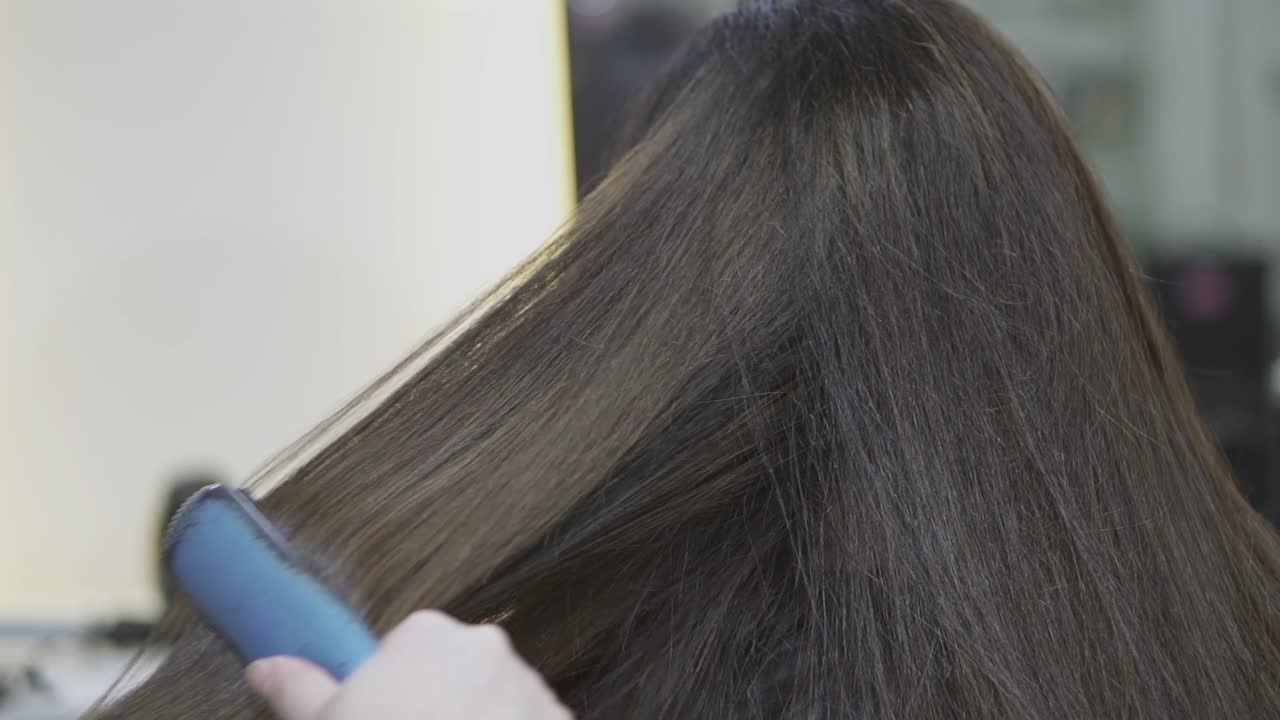 Stylisti harjaa ja suoristaa naisen ruskeita hiuksia kampaamossa keskittyen sileään muotoiluun.