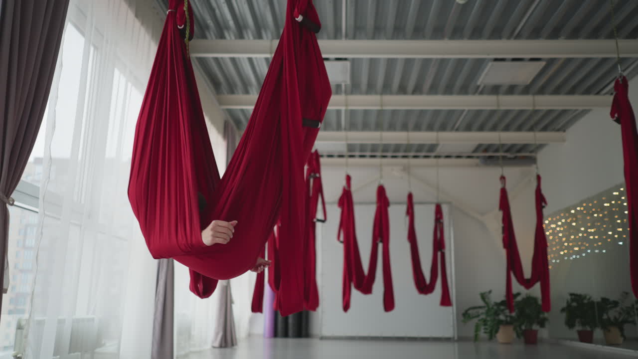 Bright yoga studio with floor to ceiling windows and sheer curtains as fitness coach lies comfortably in red silk hammock swinging gently, smiling, conveying wellness balance and calm