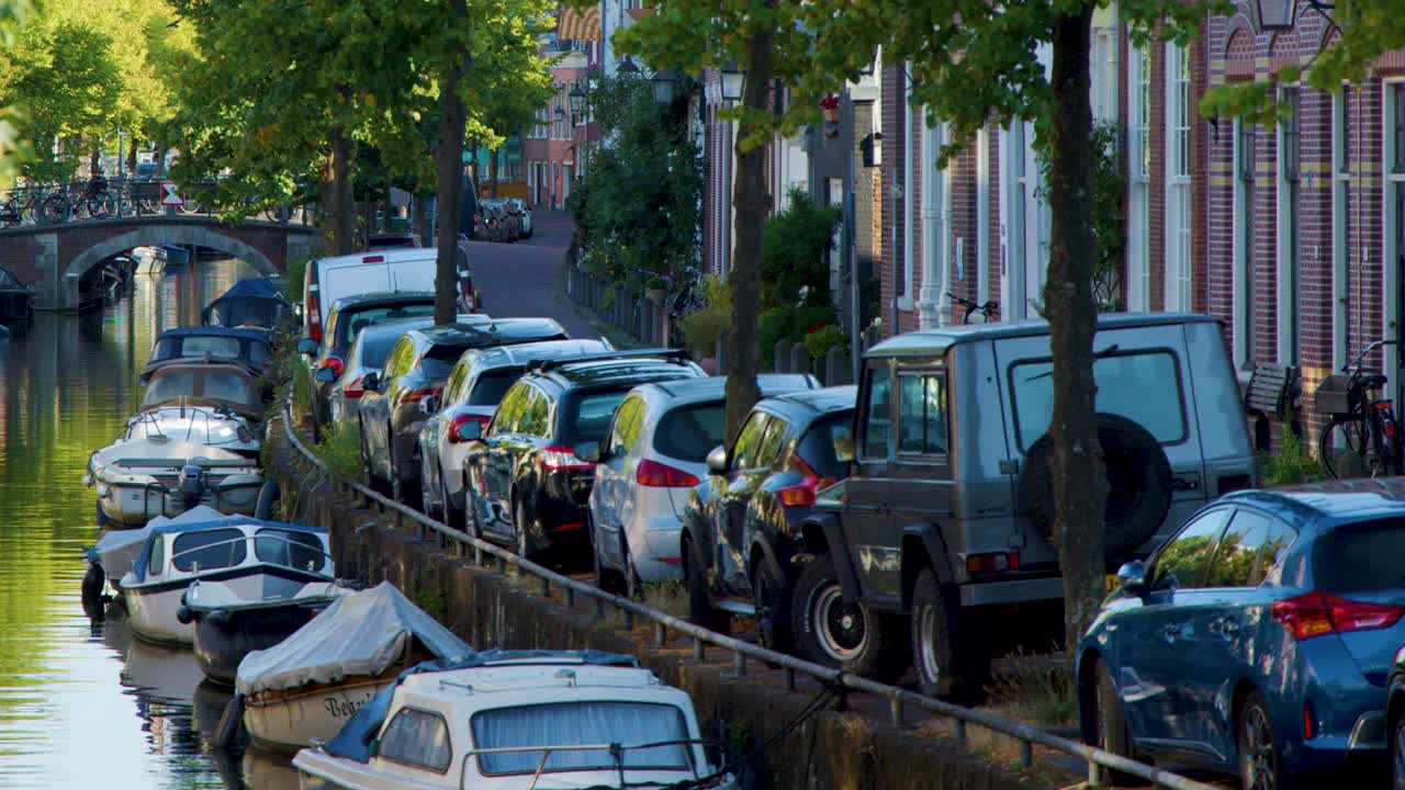 Daytime camera pan along Amsterdam canal, showing parked cars, boats, trees, and bridge