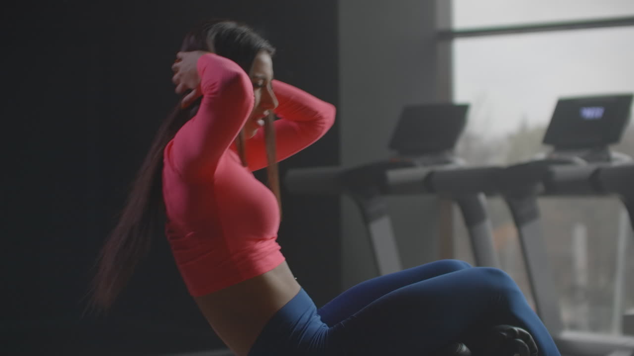 The young fit female is doing oblique abdominal muscles exercise in gym. She is controlling her breath and technique. She is changing side every time. This is a productive morning workout in the fitness center.