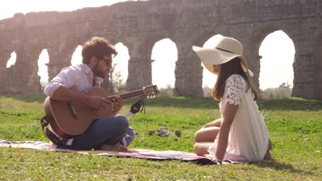 happy young lovely couple sitting lying on a blanket in the grass in front of ancient roman aqueduct ruins in parco degli acquedotti park in rome romantic play guitar sing beautiful girl with hat bucolic slow motion