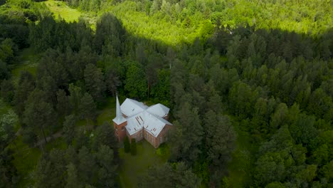 aerial pullback or revealing shot of a red stone church with a white roof in a thick forest while clouds and sun are creating shadows on the treetops during summer sunny day. alatskivi church, peipsi