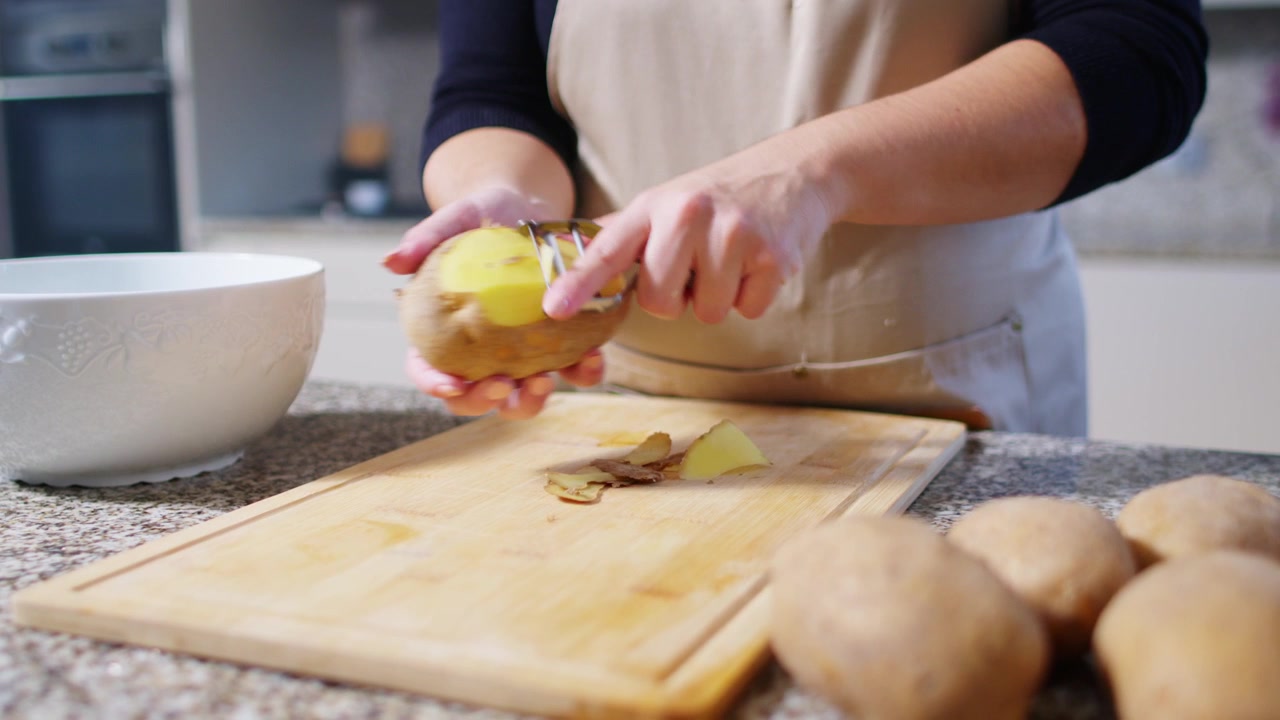 Ripresa al rallentatore di una donna che pela patate con un pelapatate, con patate fresche in primo piano, evidenziando il processo di pelatura in cucina.