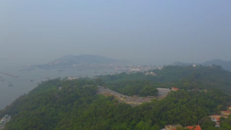 drone shot traveling backward above a forest on a hill away from a village on an island during a very polluted day.
