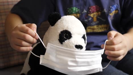 young caucasian boy puts mask on stuffed baby panda, covil-19 quarantine