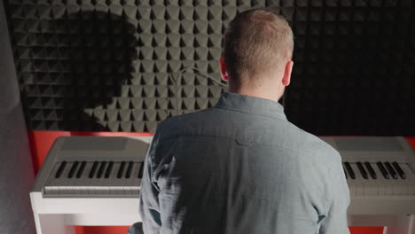 back shot of a man in a blue shirt playing a white piano, with his shadow cast against a black acoustic wall. the scene is illuminated softly, highlighting the contrast between the man, the piano