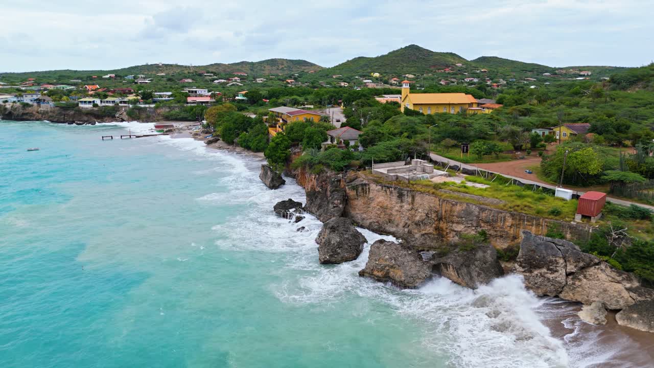 Drone che scende lungo una scogliera marina sulla costa di Curaçao, con massi nell'acqua, onde epiche che si infrangono e sabbia in sospensione
