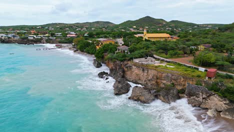 Drone-descends-along-sea-cliff-on-coastline-of-Curacao-boulders-in-water-with-epic-waves-crashing-and-suspended-sand