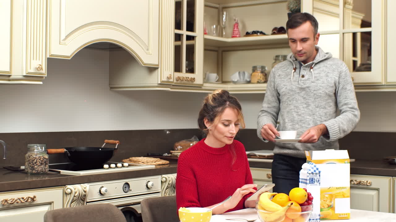 CHISINAU, MOLDOVA - SEPTEMBER 25, 2018: A smiling couple on a kitchen. Man serving woman a cup of tea. Woman looking at the phone. Table with food