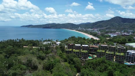 Smooth aerial pan left revealing coastline, beach, and tropical buildings among lush green hills in Phuket