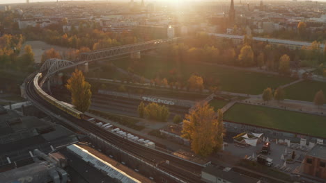 Subway-Train-from-Aerial-perspective-passing-bridge-through-public-park-in-Berlin,-Germany-in-Yellow-and-Orange-autumn-colors-and-Sunset-golden-light