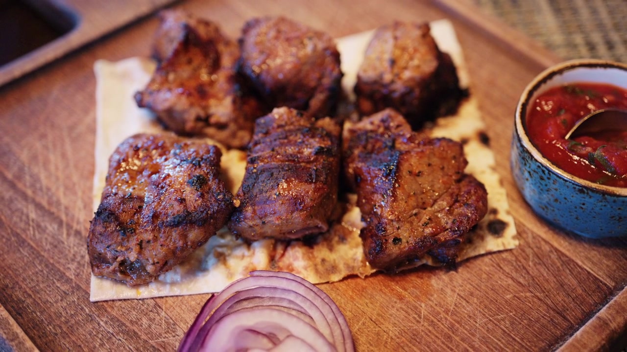 Pezzi di carne grigliata sono disposti su pane piatto, serviti con una piccola ciotola di salsa rossa e fette di cipolla rossa su una superficie di legno.