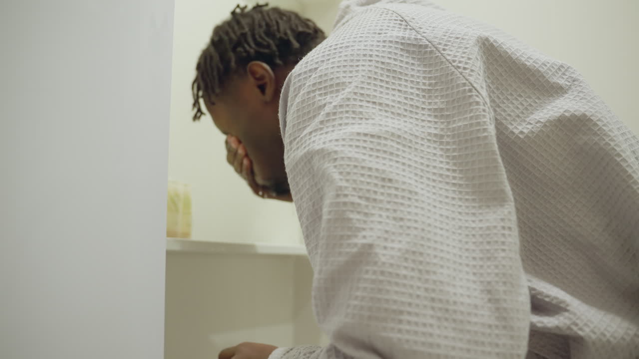 Black youth in bathrobe standing at bathroom sink, washing hands, close-up focus on hands, personal hygiene routine, modern clean bathroom, soft natural lighting, fresh start to day