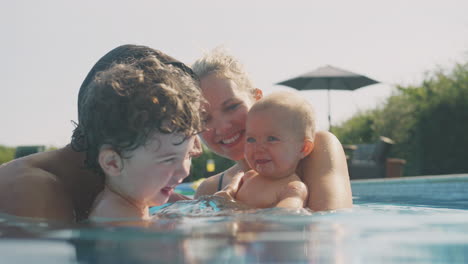 family with young son and baby having fun on summer vacation playing and splashing in swimming pool