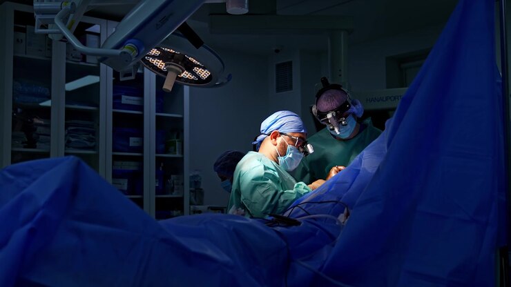 Approaching a patient covered with dark blue sheet on the operating table. Two doctors in surgical loupes perform trauma surgery.