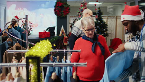 trabajador que ayuda a un cliente mayor a elegir la vestimenta adecuada para regalarle a su esposo para la próxima temporada navideña. cliente navegando a través de ropa en tienda de moda ornamentada festiva