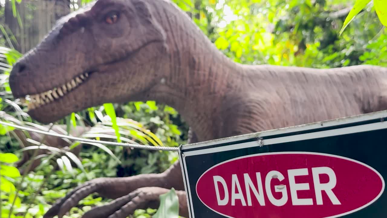 A lifelike prehistoric predator stands behind a red danger sign in a lush tropical environment