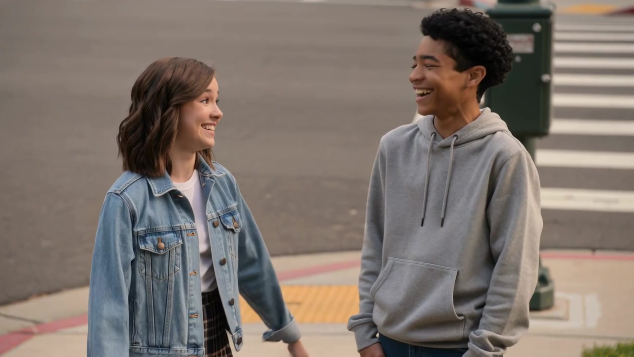 Gesturing denim jacket girl speaking gray hoodie boy sharing laughs at curb near tactile paving
